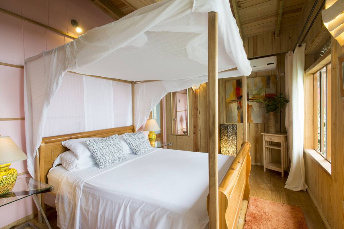 Ein hölzernes Himmelbett mit Baldachin, ein Nachttisch mit Lampe und Blick ins Zimmer.