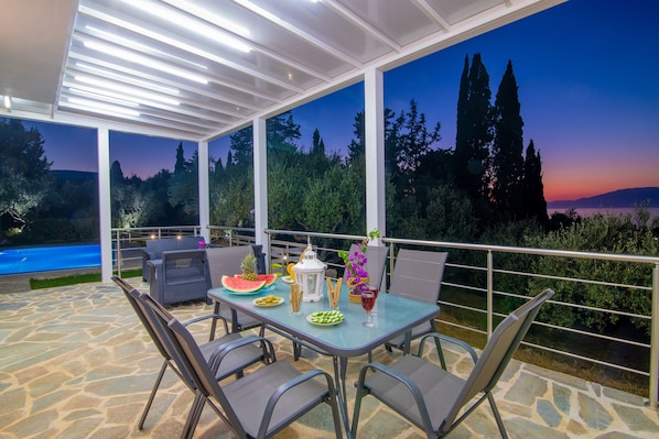 Eine Terrasse im Freien mit Esstisch und Stühlen, mit Blick auf eine schöne Landschaft aus Bäumen und einem Gewässer.