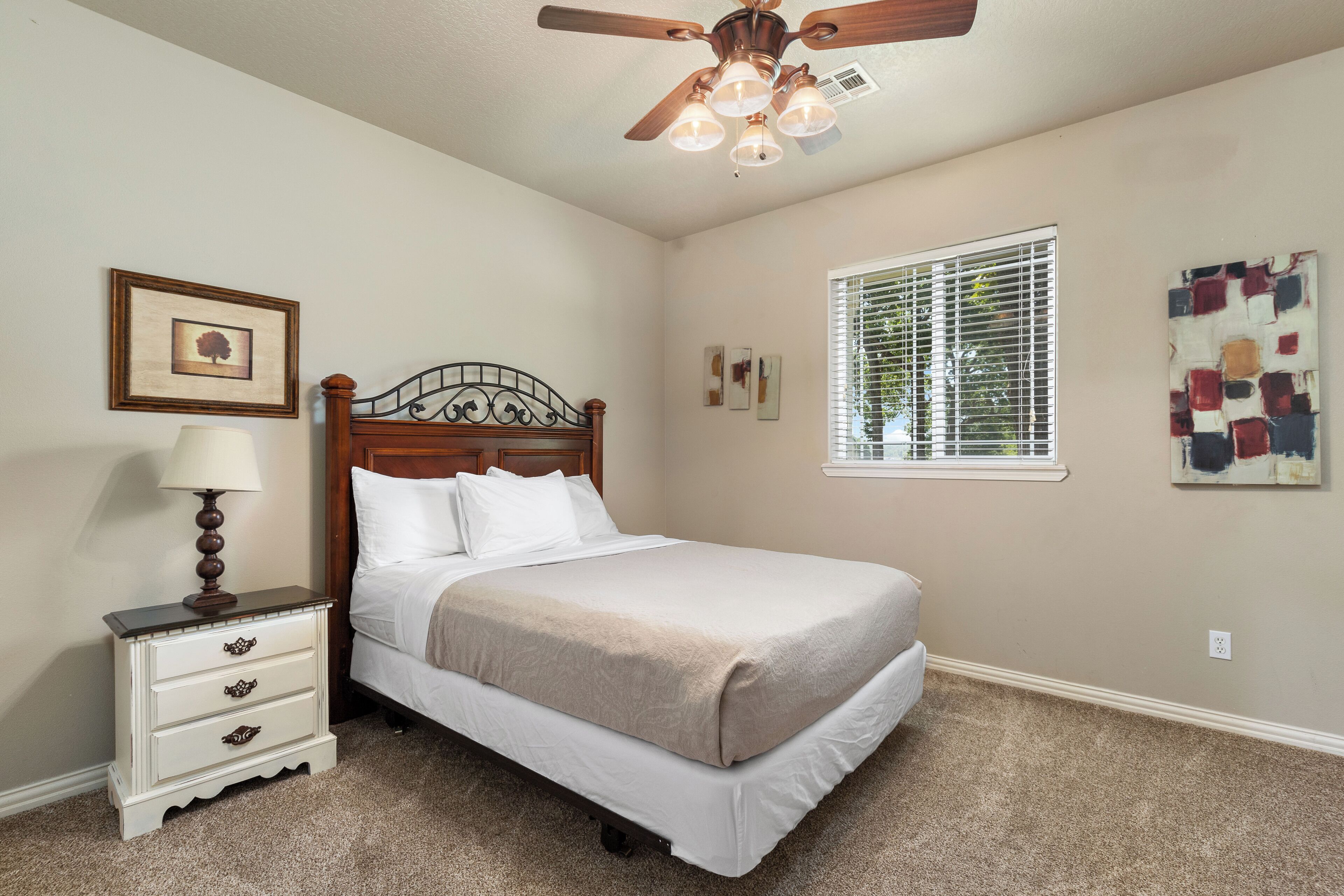 A bedroom with a bed, a nightstand, a ceiling fan, a window with blinds, and wall art.