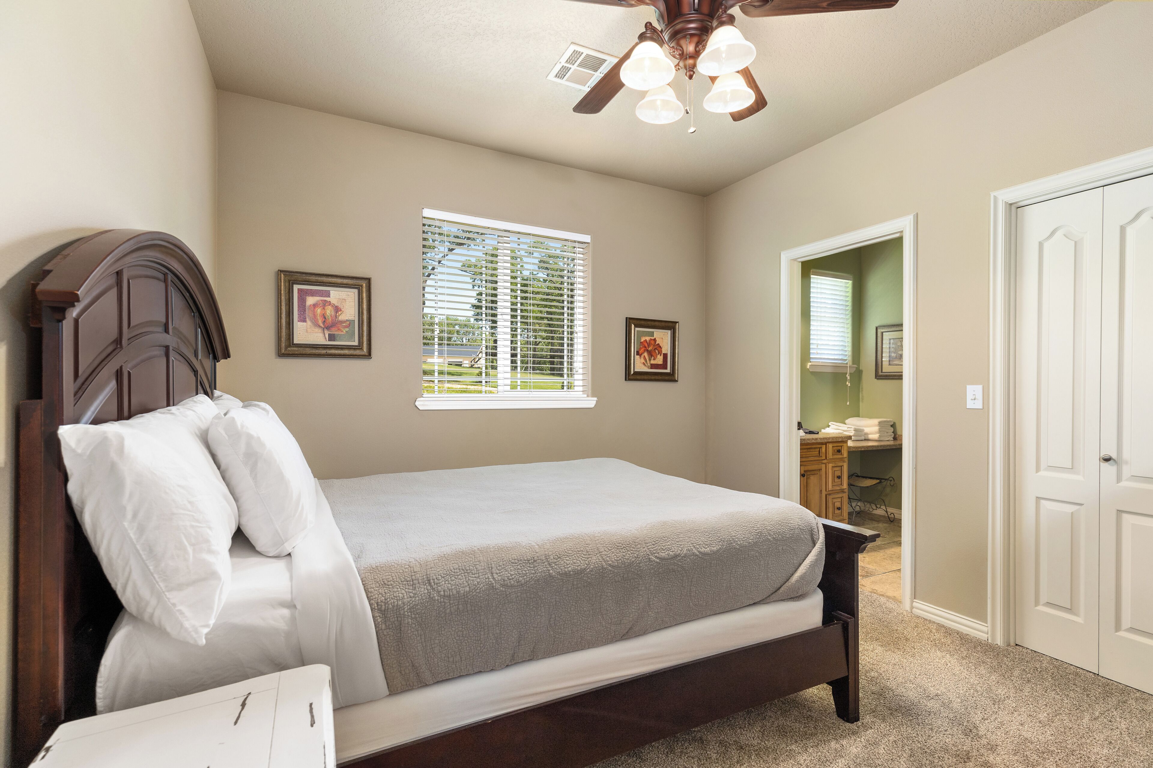 A bedroom with a bed, a ceiling fan, a window with blinds, and a door leading to a bathroom.
