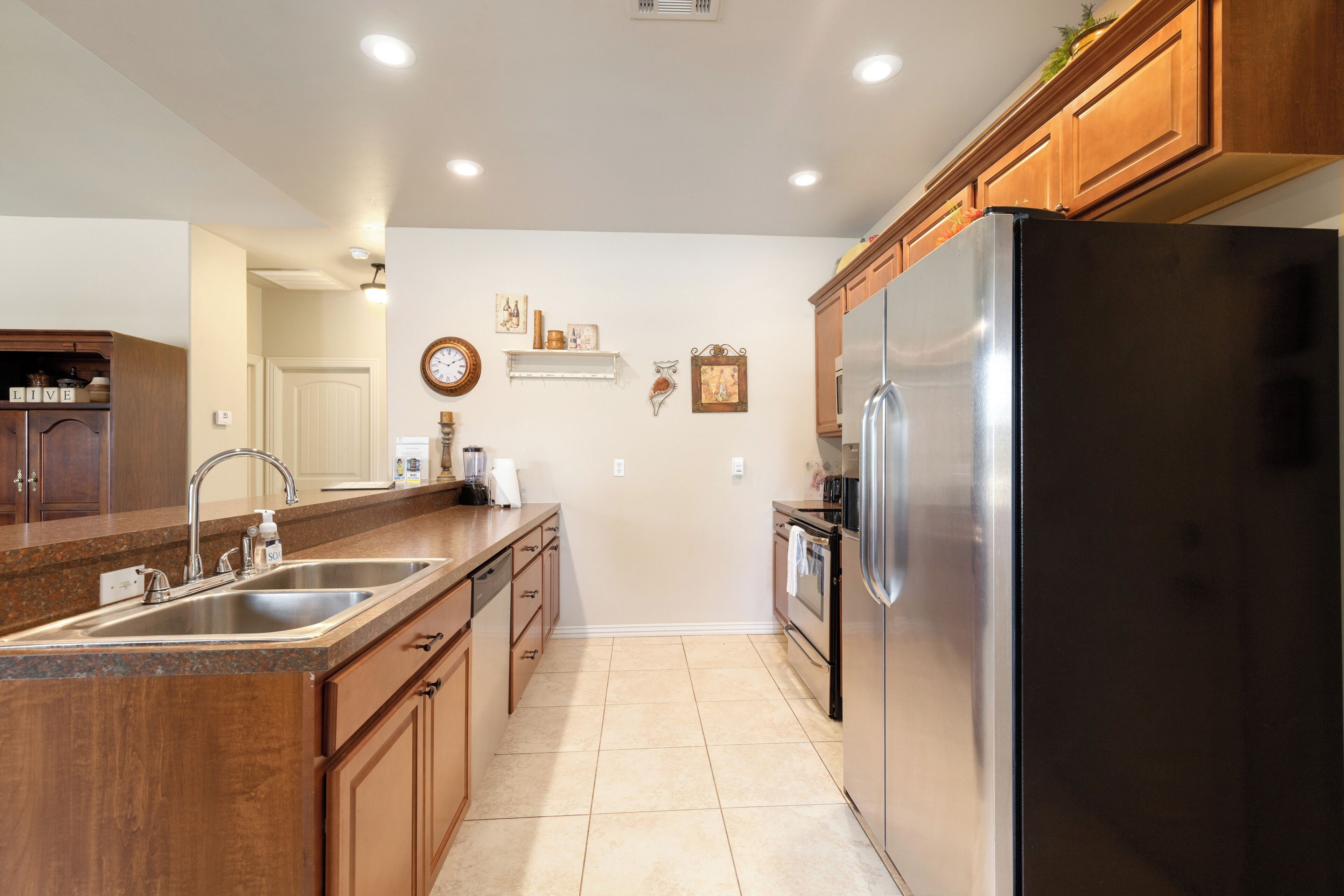 A kitchen with a stainless steel refrigerator, wooden cabinets, a double sink, and a tiled floor.