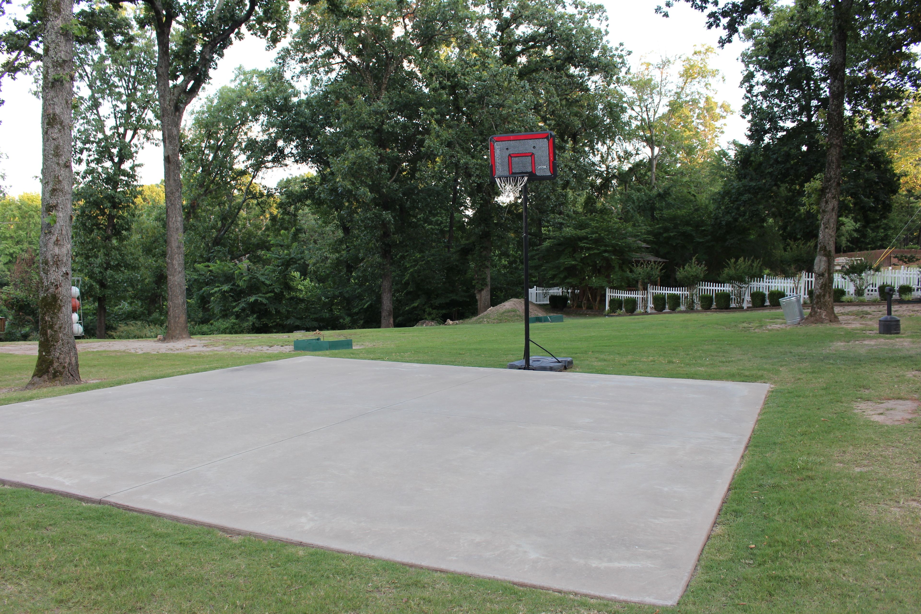 A basketball court with a hoop, surrounded by trees and a white fence.