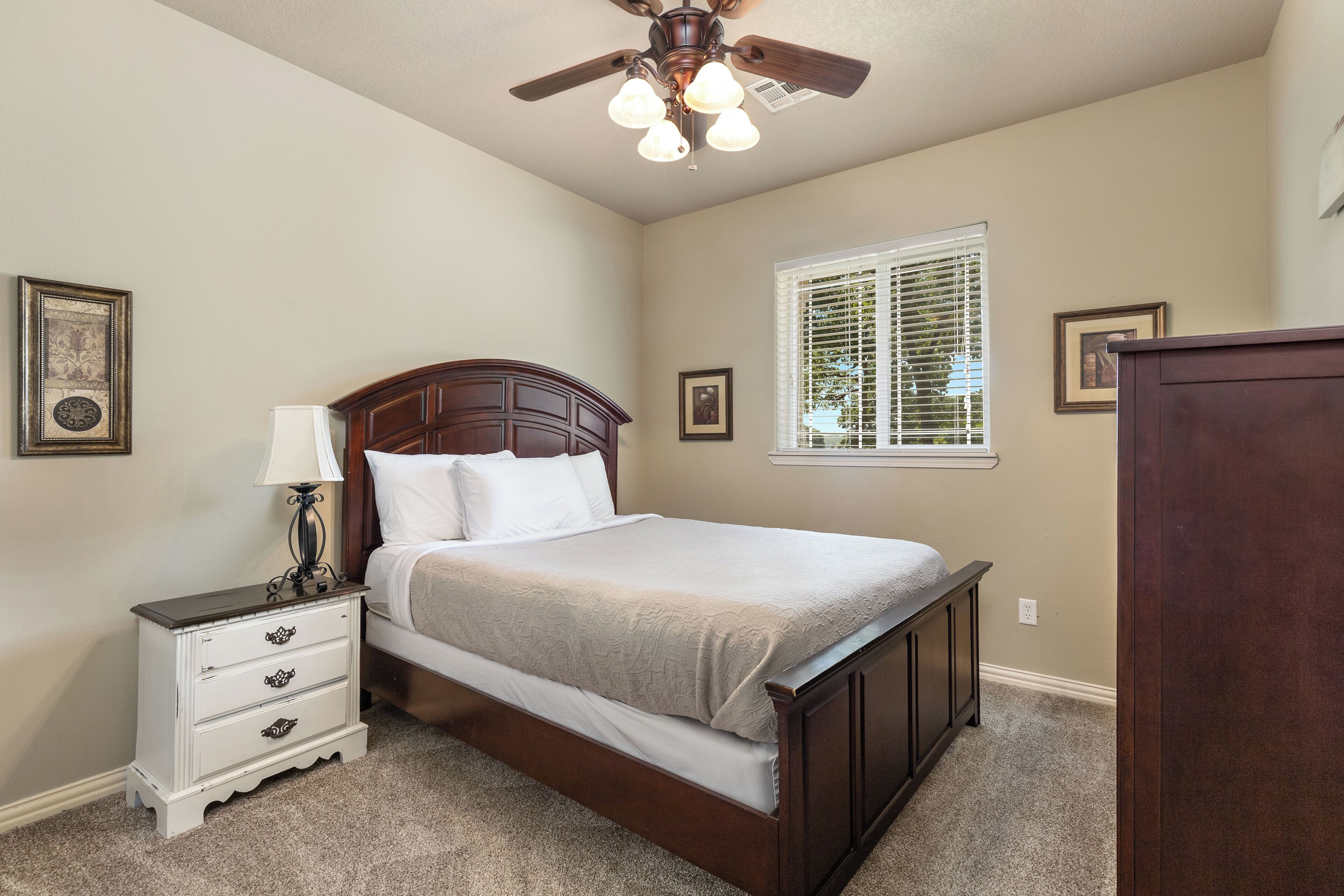 A bedroom with a bed, nightstand, lamp, ceiling fan, window, and wall decorations.