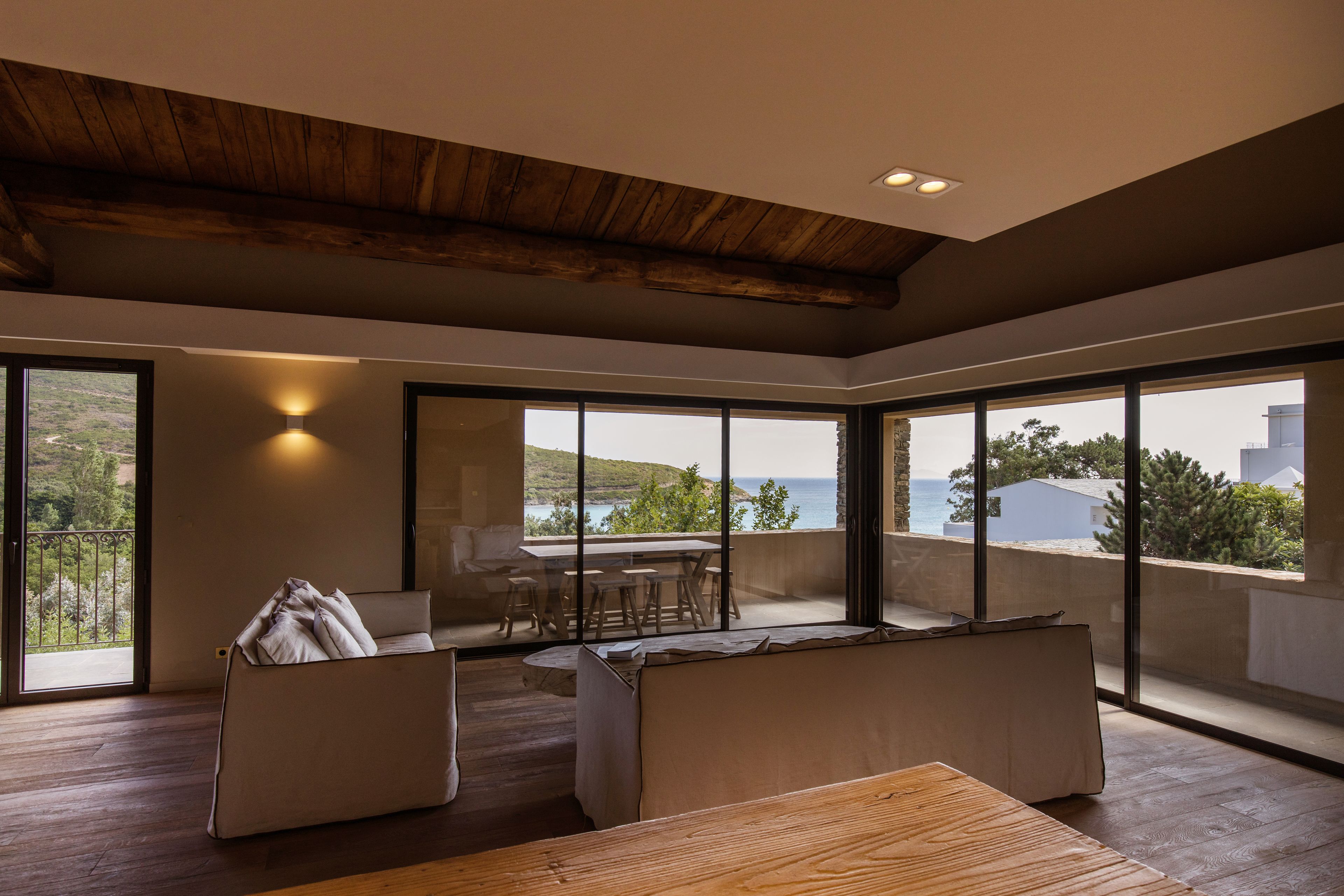 Un salon moderne avec un plafond en bois, de grandes fenêtres, un canapé, une table et un balcon offrant une vue sur la mer.