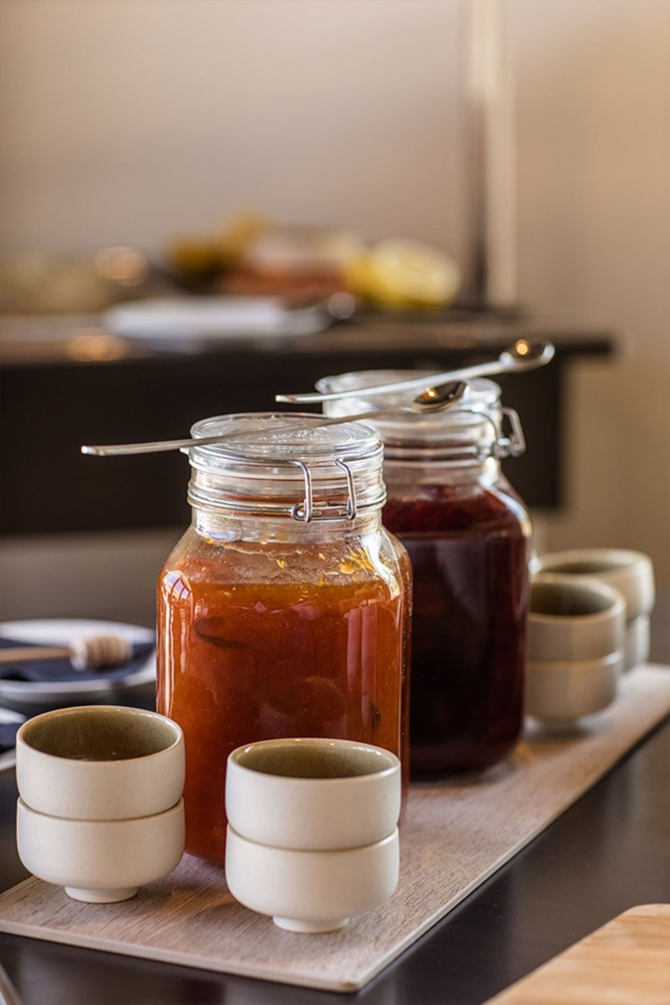 Two glass jars with metal lids and a spoon, placed on a wooden tray with two small ceramic cups.