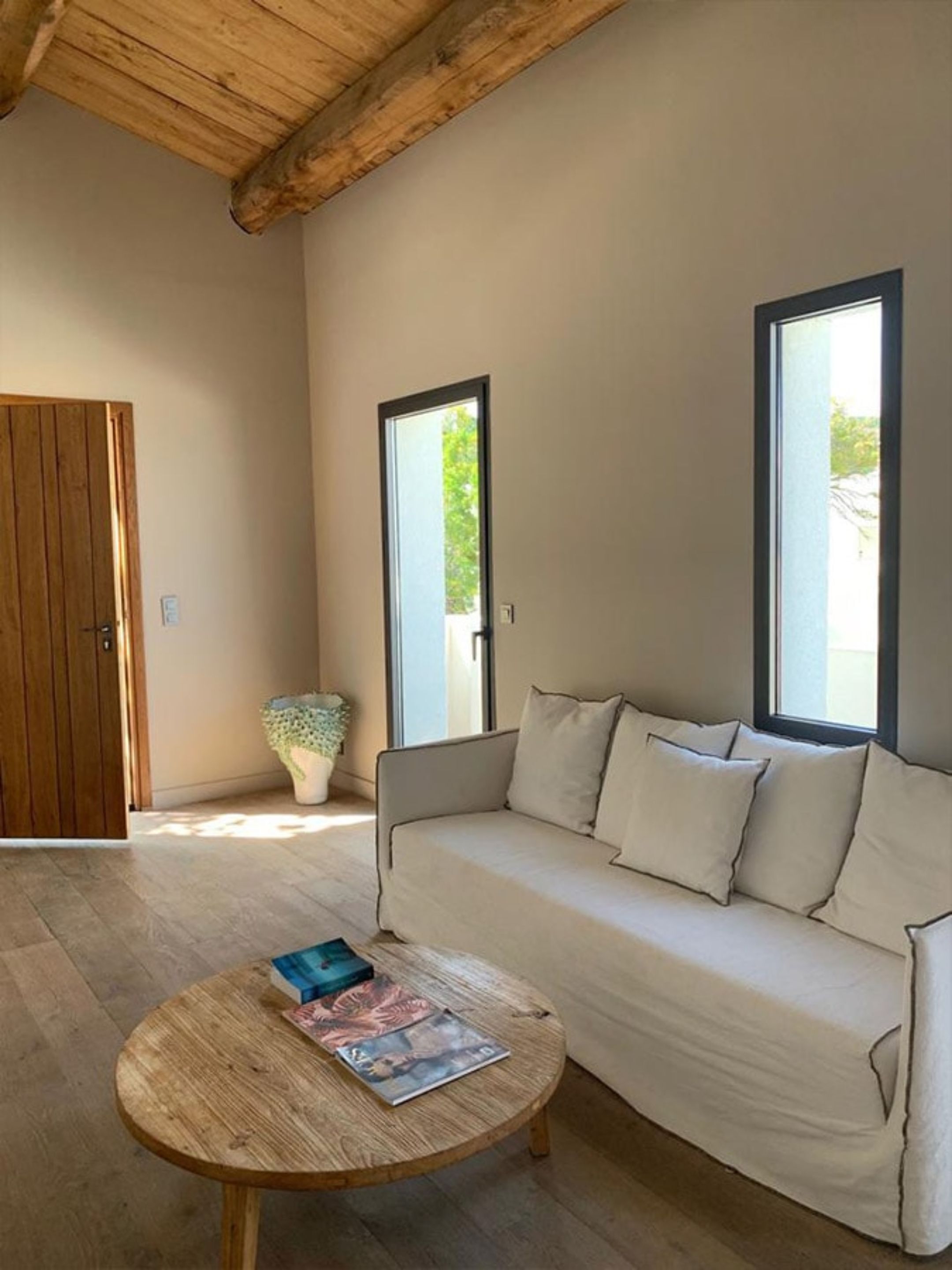 Une pièce avec un plafond en bois, un canapé beige agrémenté de coussins, une table basse en bois sur laquelle sont posés des magazines, et deux fenêtres équipées de stores.