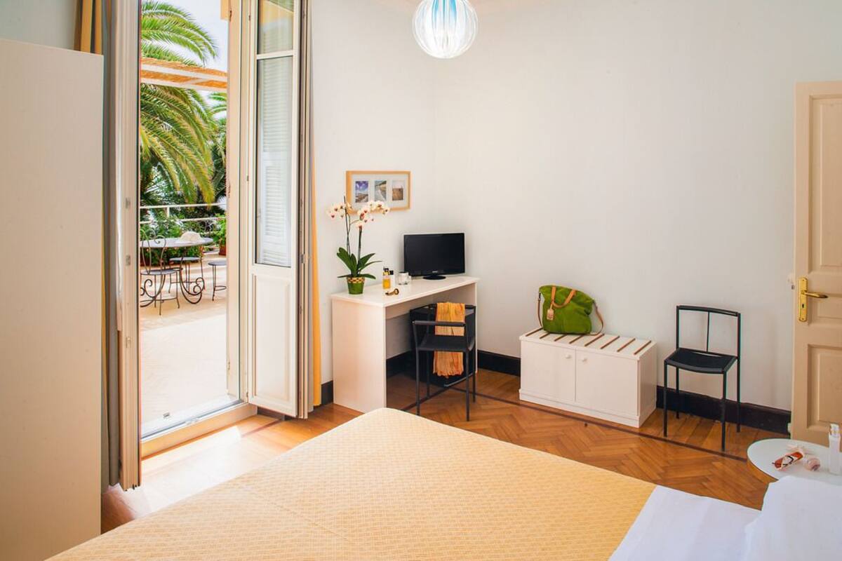 A hotel room with a bed, a desk with a computer, a chair, a small table with a bag, and a view of the outdoors.