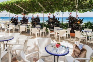 An outdoor café with white wicker chairs and tables, some with blue accents, arranged on a stone patio overlooking the sea. - Hotel Beau Sejour (Alassio)