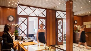 A reception area with wooden paneling, a reception desk, a clock, and a glass partition. - Hotel Beau Sejour (Alassio)