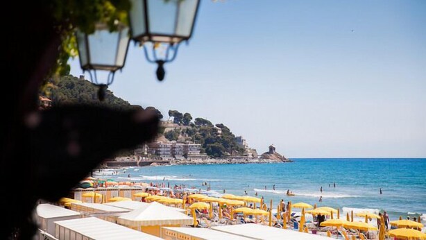 A beach with rows of yellow umbrellas and white tents, a clear blue sky, and a hilly landscape in the background. - Hotel Beau Sejour (Alassio)