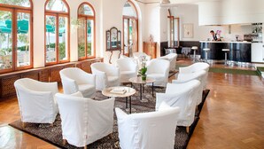 A spacious lounge area with white upholstered chairs and a central round table, large arched windows, and a view of the sea. - Hotel Beau Sejour (Alassio)