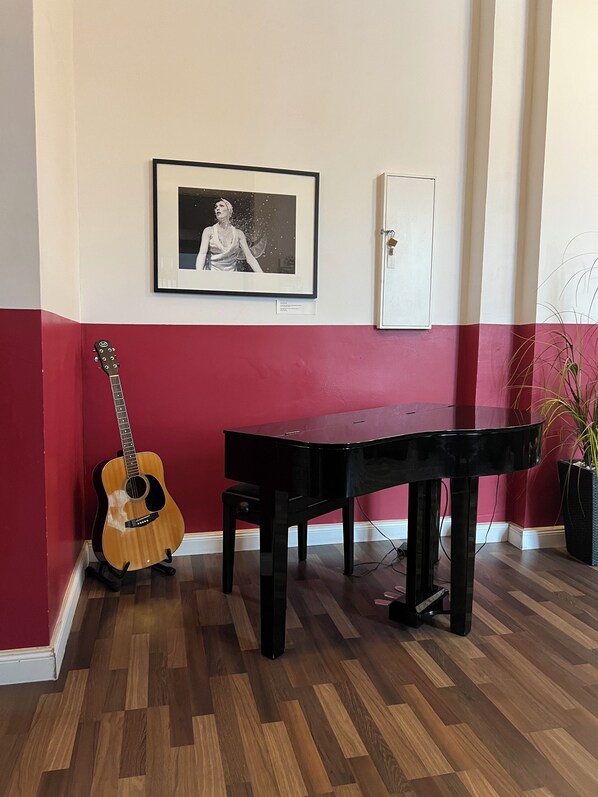 Una habitación con piso de madera, un piano negro, una guitarra apoyada contra una pared roja y un cuadro en la pared.