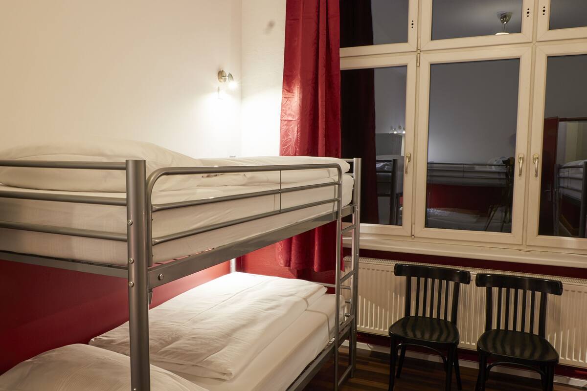 A bunk bed with a white mattress and metal frame, two black chairs, a window with red curtains, and a radiator below.