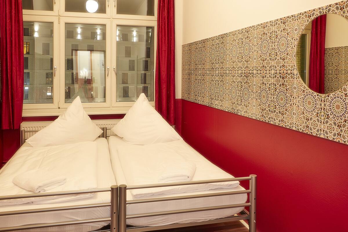 A double bed with white linens, a window with red curtains, and a decorative wall with a circular mirror.