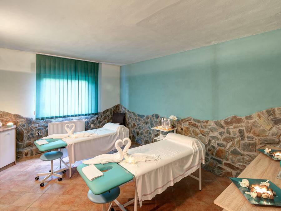 Une salle de spa comprenant deux tables de massage, un mur en pierre, une fenêtre avec des rideaux et un petit meuble.