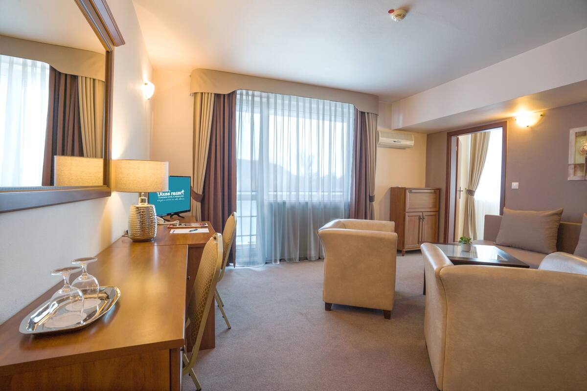 Ein Hotelzimmer mit Schreibtisch, zwei Sesseln, einer Couch, einem Essbereich mit Stuhl, einem Fernseher und einem großen Fenster mit Vorhängen.