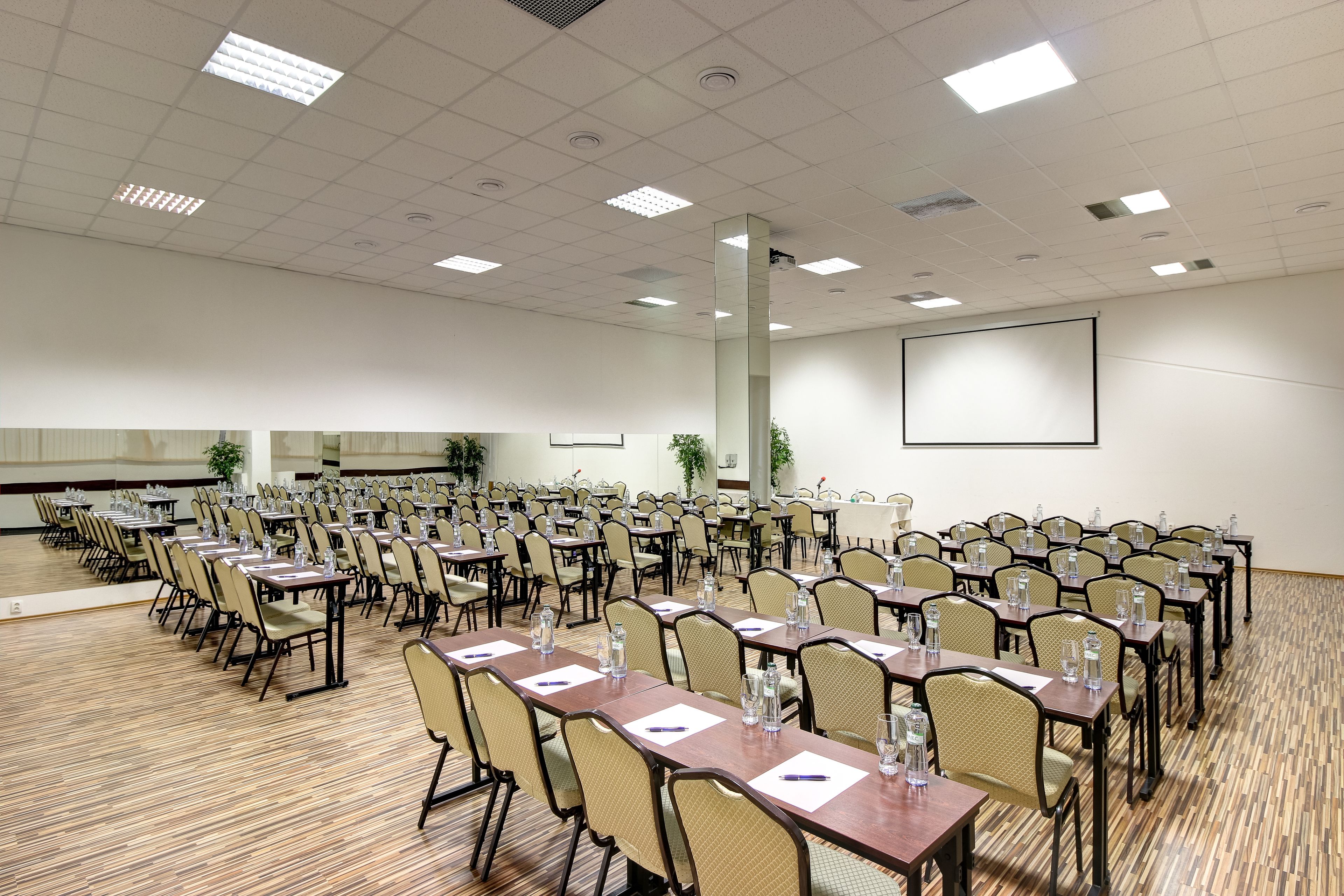 A conference room with rows of tables and chairs, a large screen, and a wooden floor.