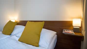 Uma cama bem arrumada com lençóis brancos e duas almofadas amarelo-mostarda, uma cabeceira de madeira e uma mesa de cabeceira com uma luminária e uma revista.