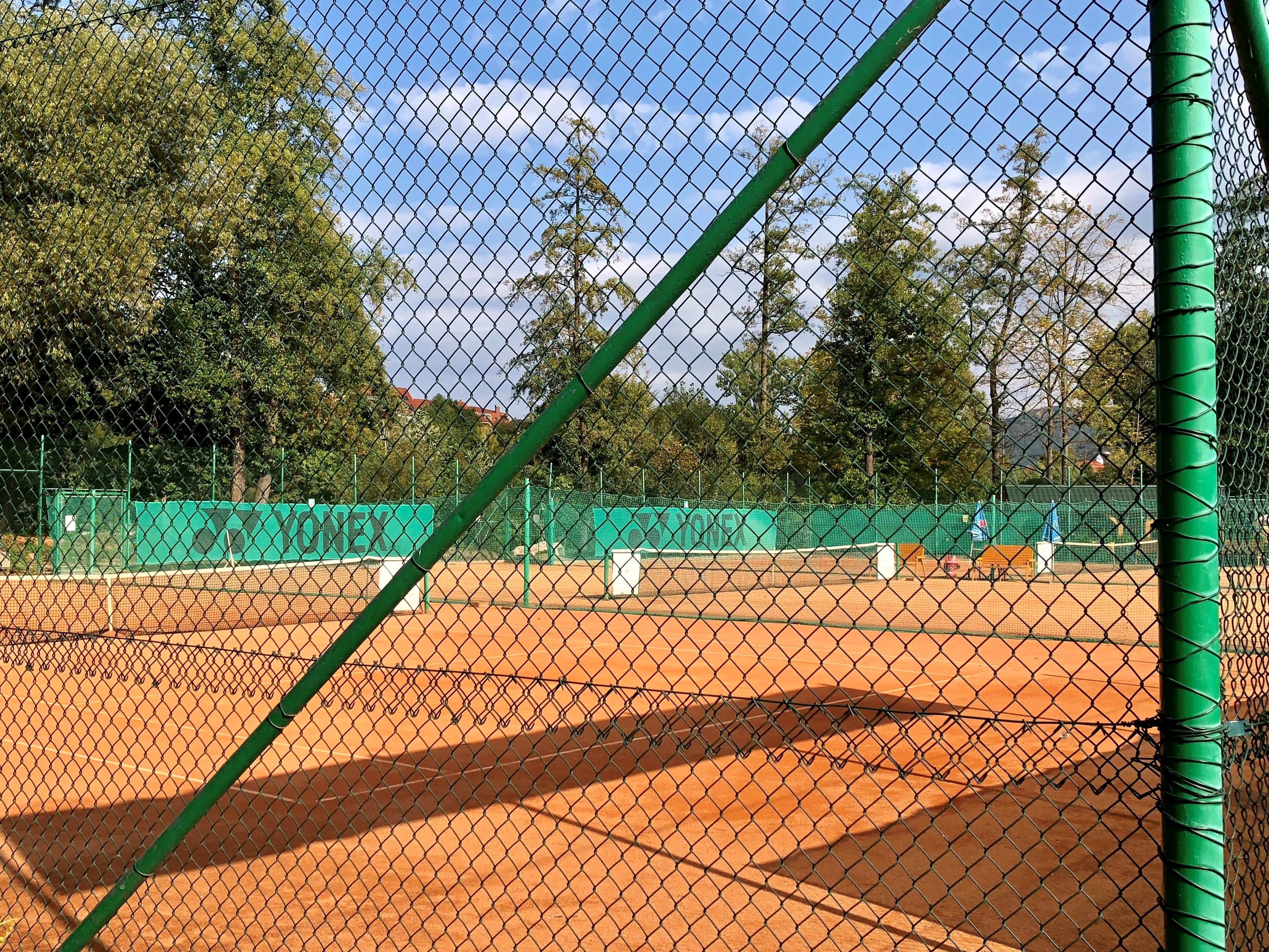 A tennis court with a chain-link fence, green netting, and a tennis ball machine.