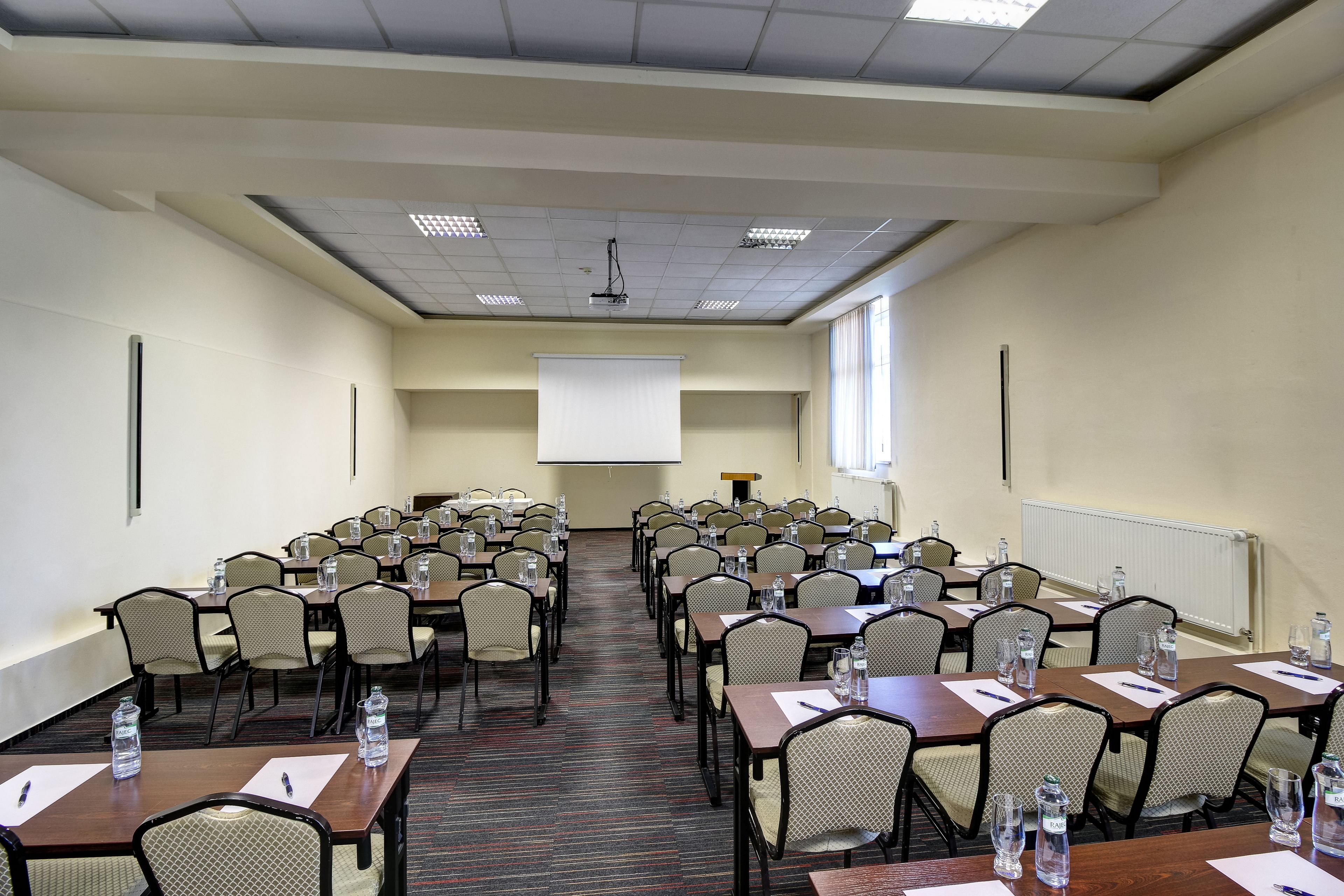 A conference room with multiple tables and chairs, equipped with water bottles and pens, and a projector screen at the front.