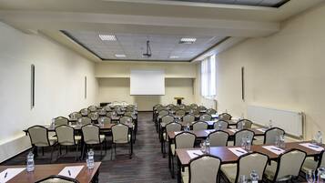 A conference room with multiple tables and chairs, equipped with water bottles and pens, and a projector screen at the front.