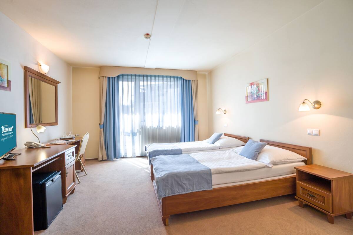 Ein Hotelzimmer mit zwei Betten, einem Schreibtisch mit Computer, einem Stuhl, einer kleinen Ablage und einem Fenster mit Vorhängen.
