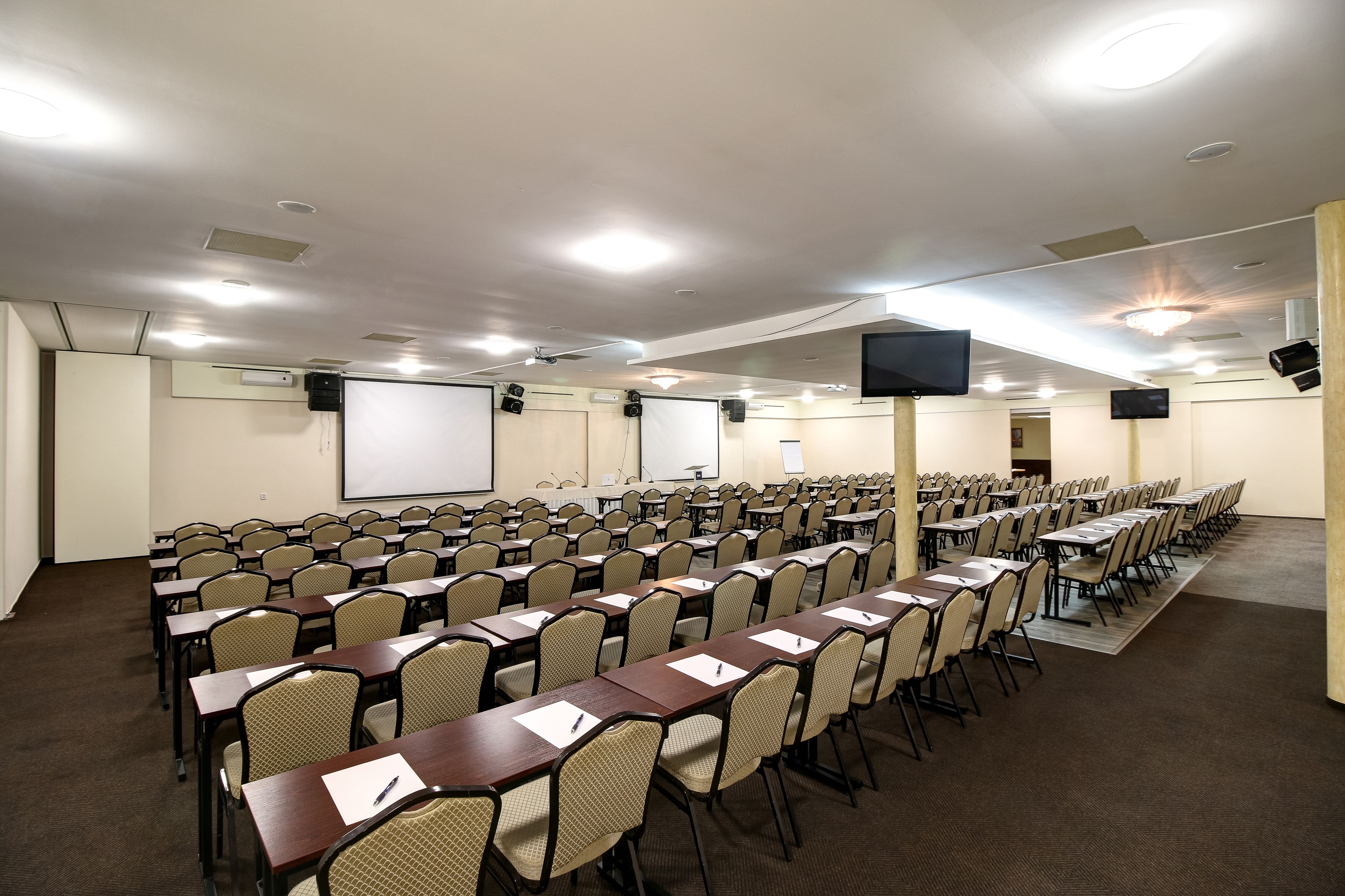A conference room with rows of tables and chairs, a projector screen, and a flat-screen TV.