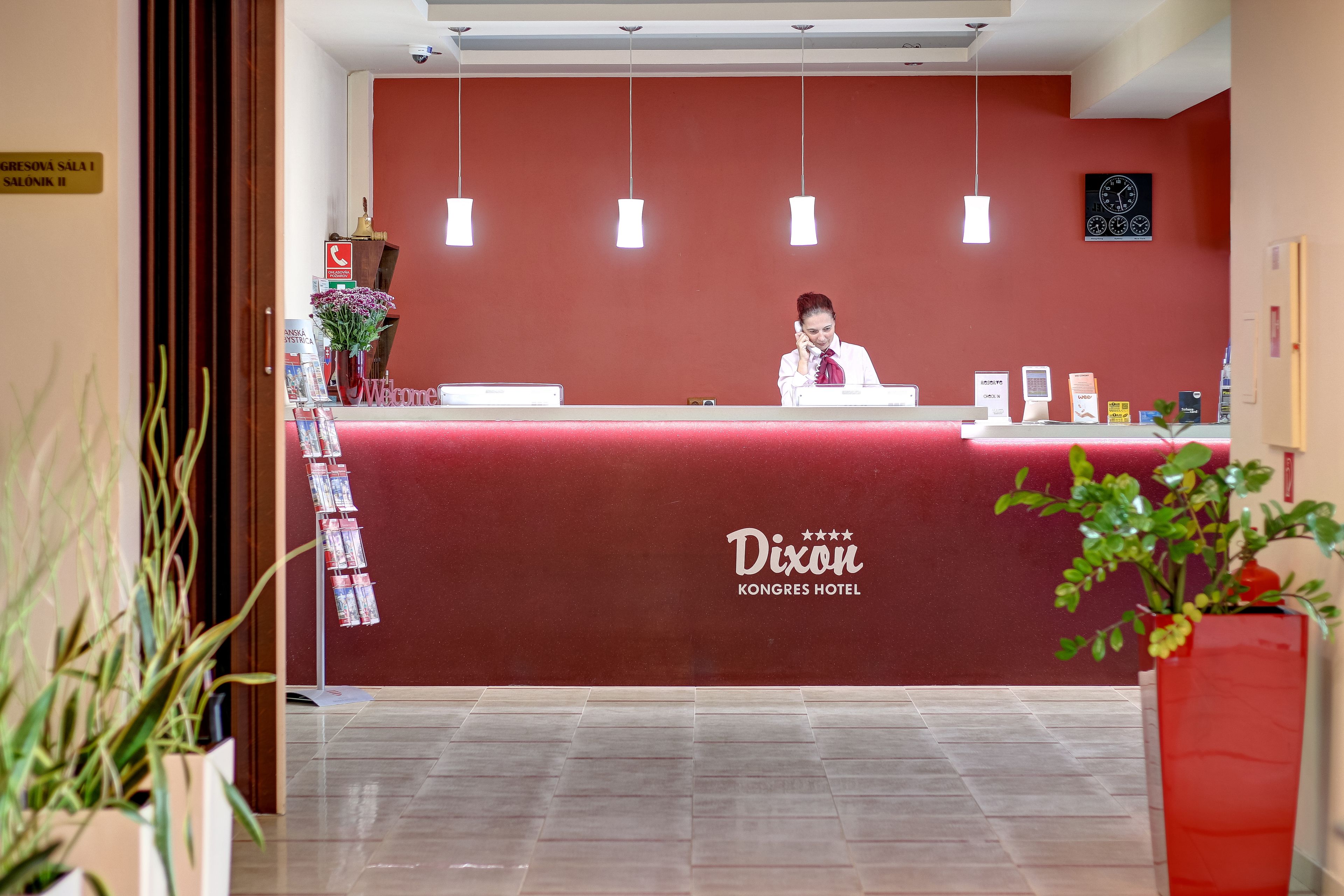 Un comptoir d’accueil à l’hôtel Dixon Kongres, avec un mur rouge, des luminaires suspendus et une horloge.