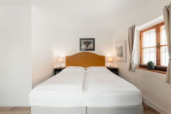 Una cama doble con sábanas blancas, dos mesitas de noche con lámparas, un cuadro enmarcado sobre la cama, una ventana con cortinas y una planta pequeña en el alféizar.