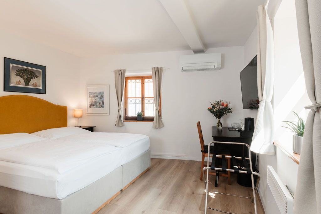 A hotel room with a bed, a desk, a chair, a window with curtains, and a wall-mounted air conditioner.