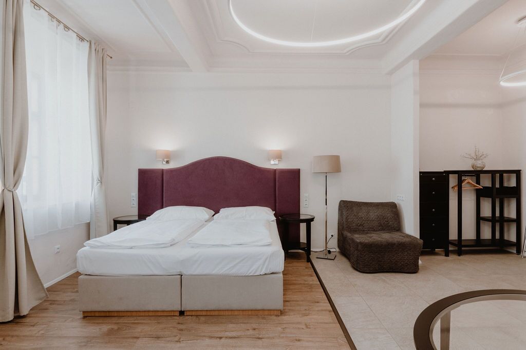 A modern hotel room with a double bed, two bedside tables, a chair, a small side table, and a wardrobe.