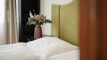 A neatly made bed with white linens, a green upholstered headboard, and a vase with flowers on a bedside table.
