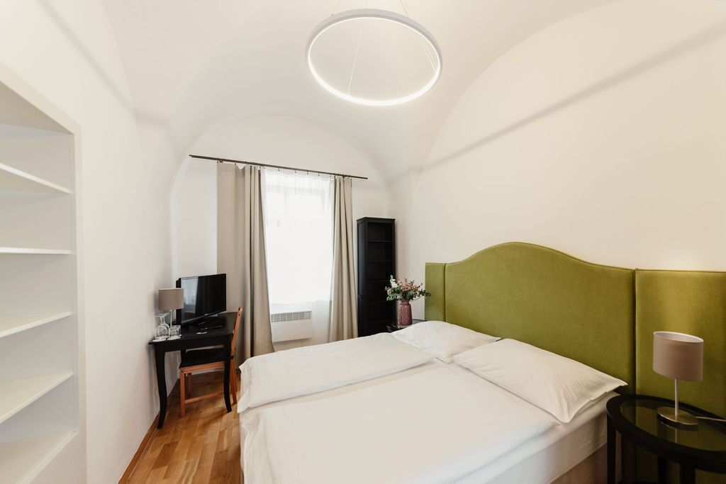 A hotel room with a bed, a desk with a computer, a chair, a TV, and a lamp.
