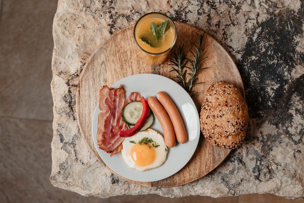 A breakfast plate with sausages, bacon, a fried egg, and a slice of bread on a wooden board.