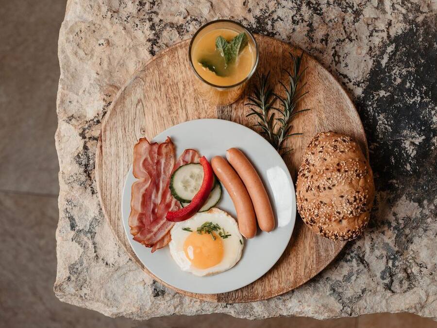 A breakfast plate with sausages, bacon, a fried egg, and a slice of bread on a wooden board.