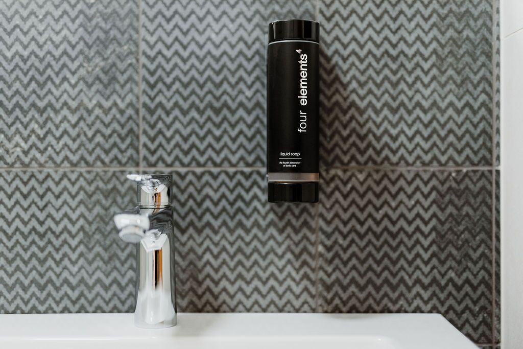 Un baño de diseño moderno, con un grifo cromado y una botella negra de jabón líquido etiquetada como "four elements".