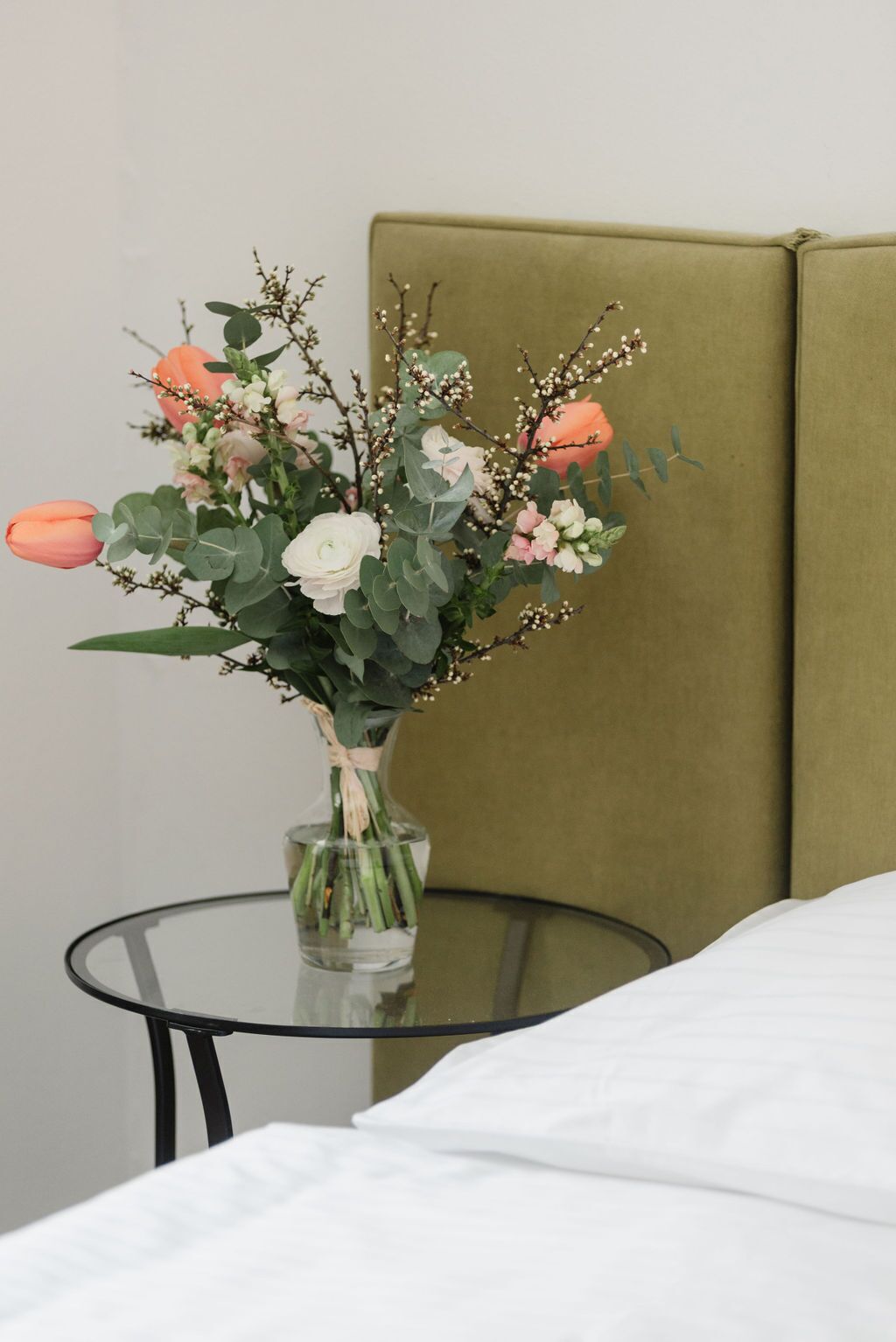 Un letto con lenzuola bianche, un tavolino di vetro con un vaso di fiori e una testiera imbottita verde.