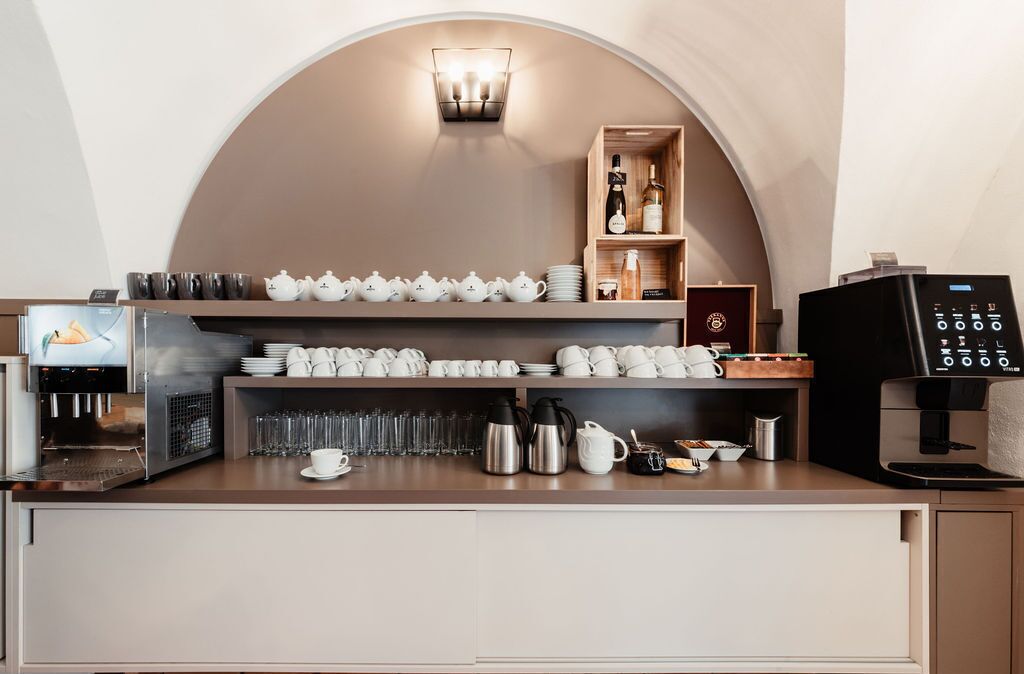 Una estación de café con una máquina de café, tazas, teteras y una repisa con botellas y vasos.