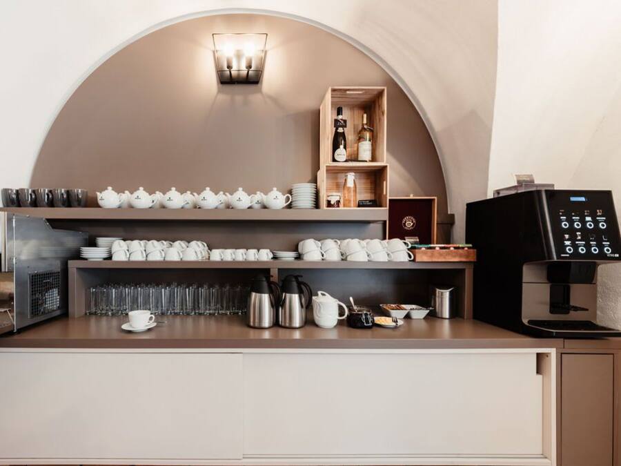 A coffee station with a coffee machine, cups, teapots, and a shelf with bottles and glasses.