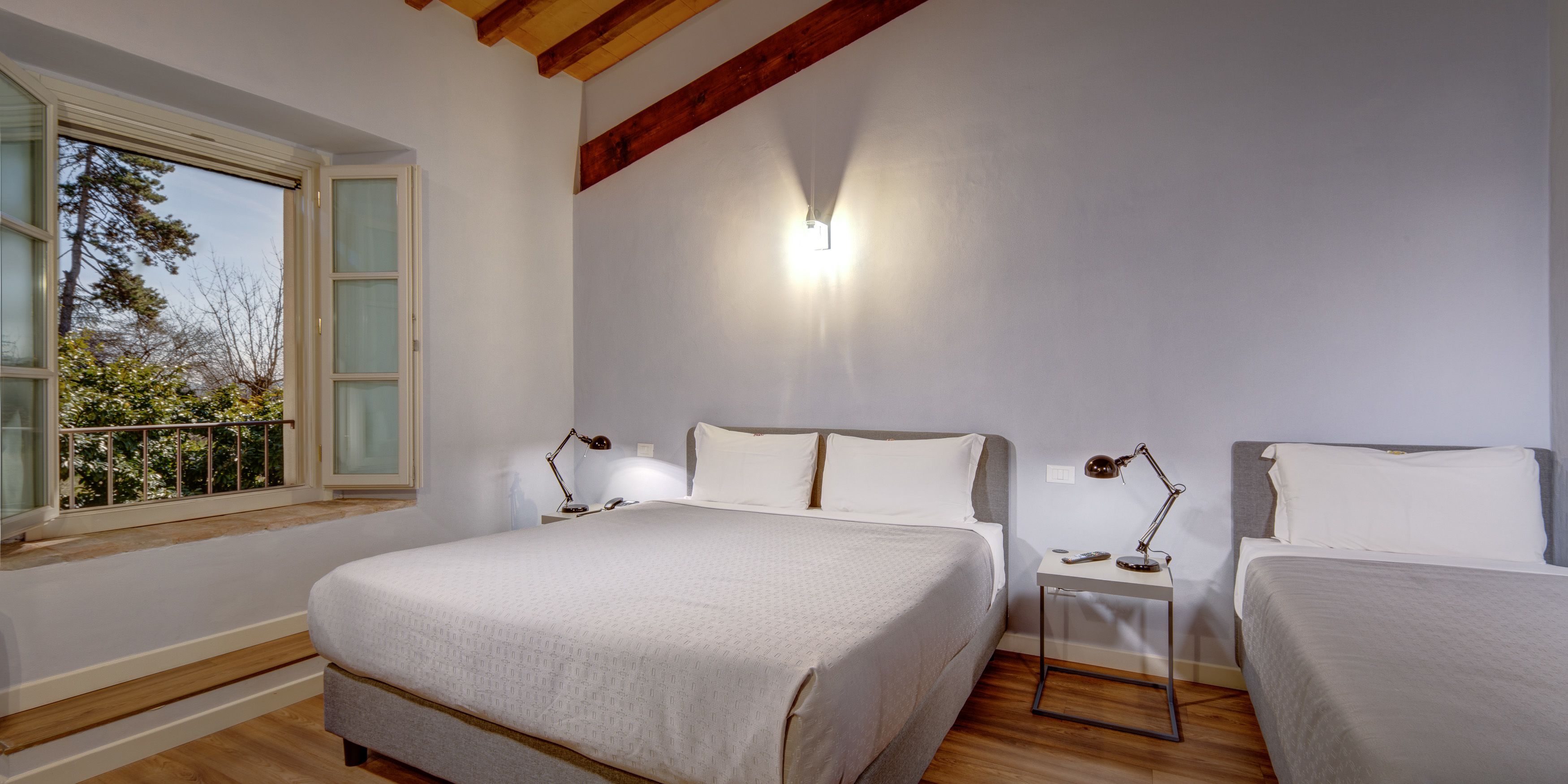 A bedroom with a large bed, two bedside tables with lamps, a window with a view of trees, and a wooden ceiling beam.