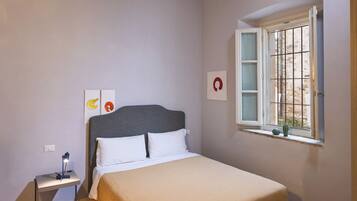 Ein Schlafzimmer mit einem Bett, einem Nachttisch mit Lampe, einem Fenster mit Blick nach draußen und einem Telefon auf einem Schreibtisch.