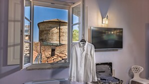 Une pièce dotée d’une grande fenêtre donnant sur un bâtiment historique, une chemise blanche accrochée au mur, une valise posée sur une petite table et un téléviseur à écran plat fixé au-dessus de la table.