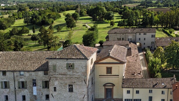 Un bâtiment historique doté d’une cour intérieure, d’un espace de stationnement avec des voitures, et offrant une vue sur un paysage verdoyant, avec des arbres et des montagnes à l’horizon.