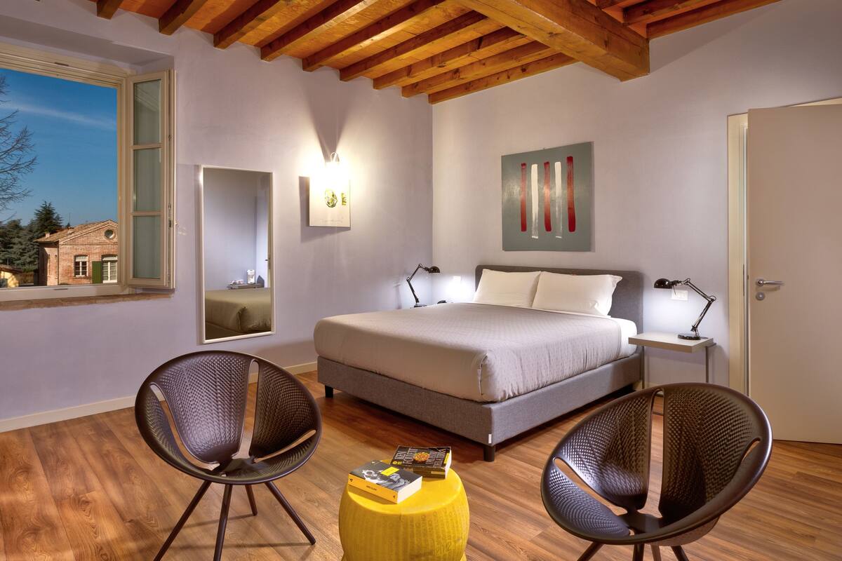 Une chambre à coucher comprenant un lit, deux chaises, une table de chevet, un miroir et une fenêtre donnant sur une maison et des arbres.