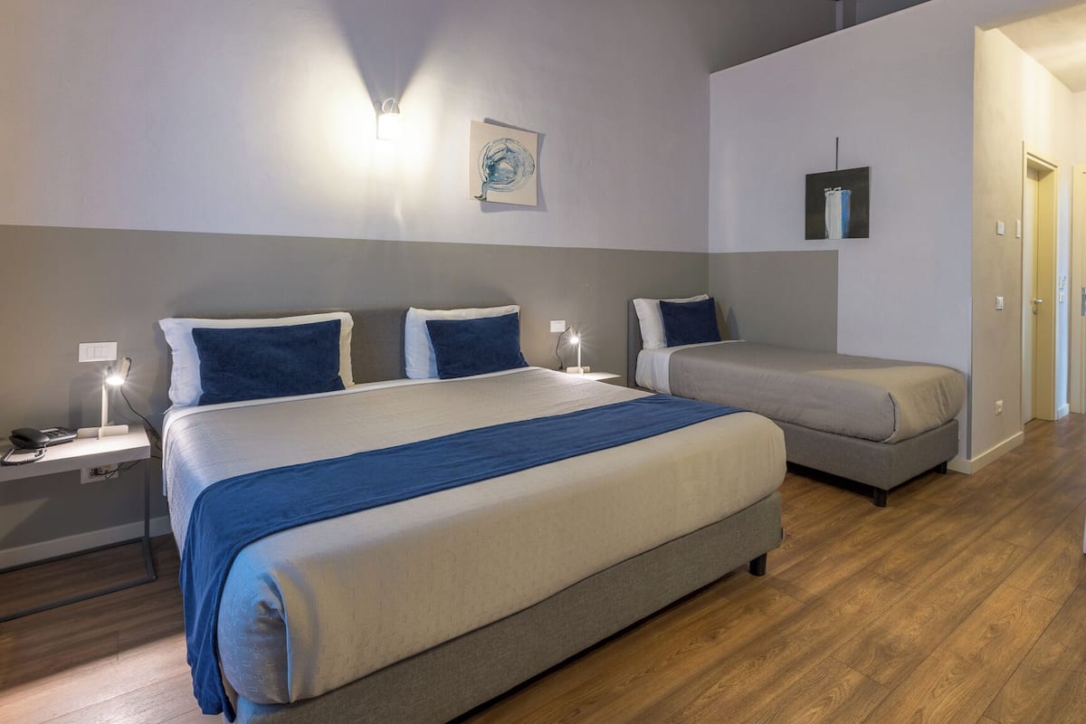 Une chambre d’hôtel avec un grand lit, un lit plus petit, un bureau et une chaise. Des lampes murales et des œuvres d’art y sont disposées.
