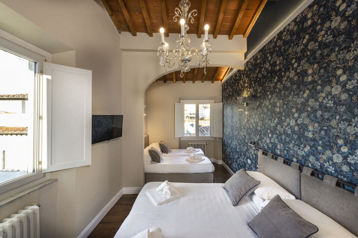 A hotel room with two beds, a chandelier, a window with a view, and floral wallpaper.