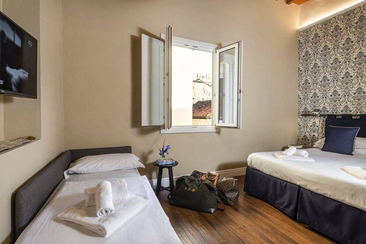 A hotel room with two beds, a television, a window with an open view, a small table with a bag, and a patterned wall.