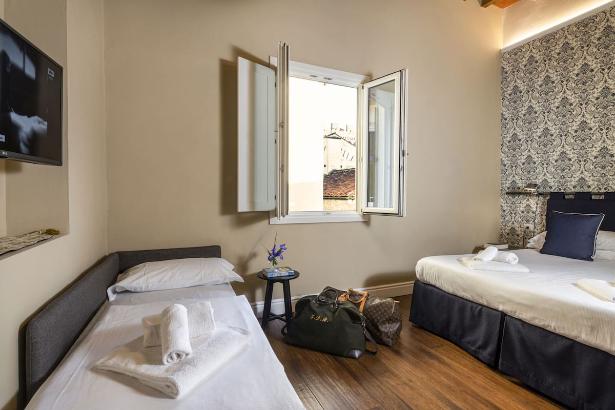 A hotel room with two beds, a TV, a window with an open view, a small table with a vase, and a bag on the floor.