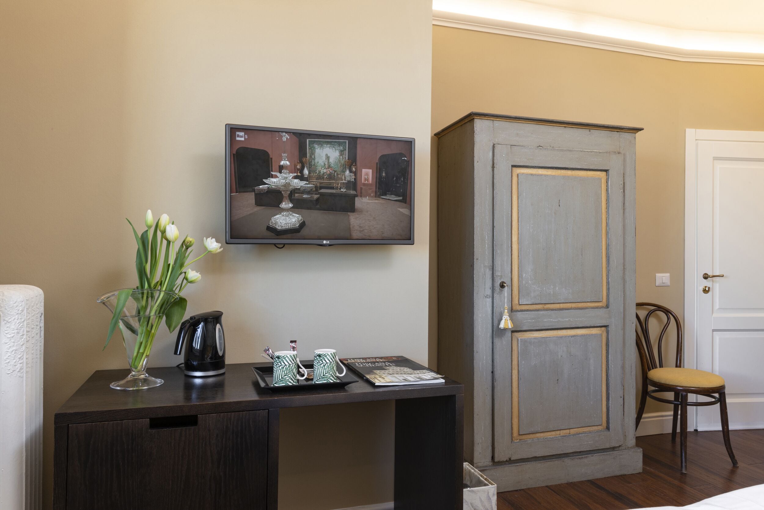 Ein Zimmer mit Fernseher an der Wand, einem Schreibtisch mit Blumen in einer Vase, einem Wasserkocher und einer Zeitschrift. Im Zimmer befindet sich ein Schrank und ein Stuhl.