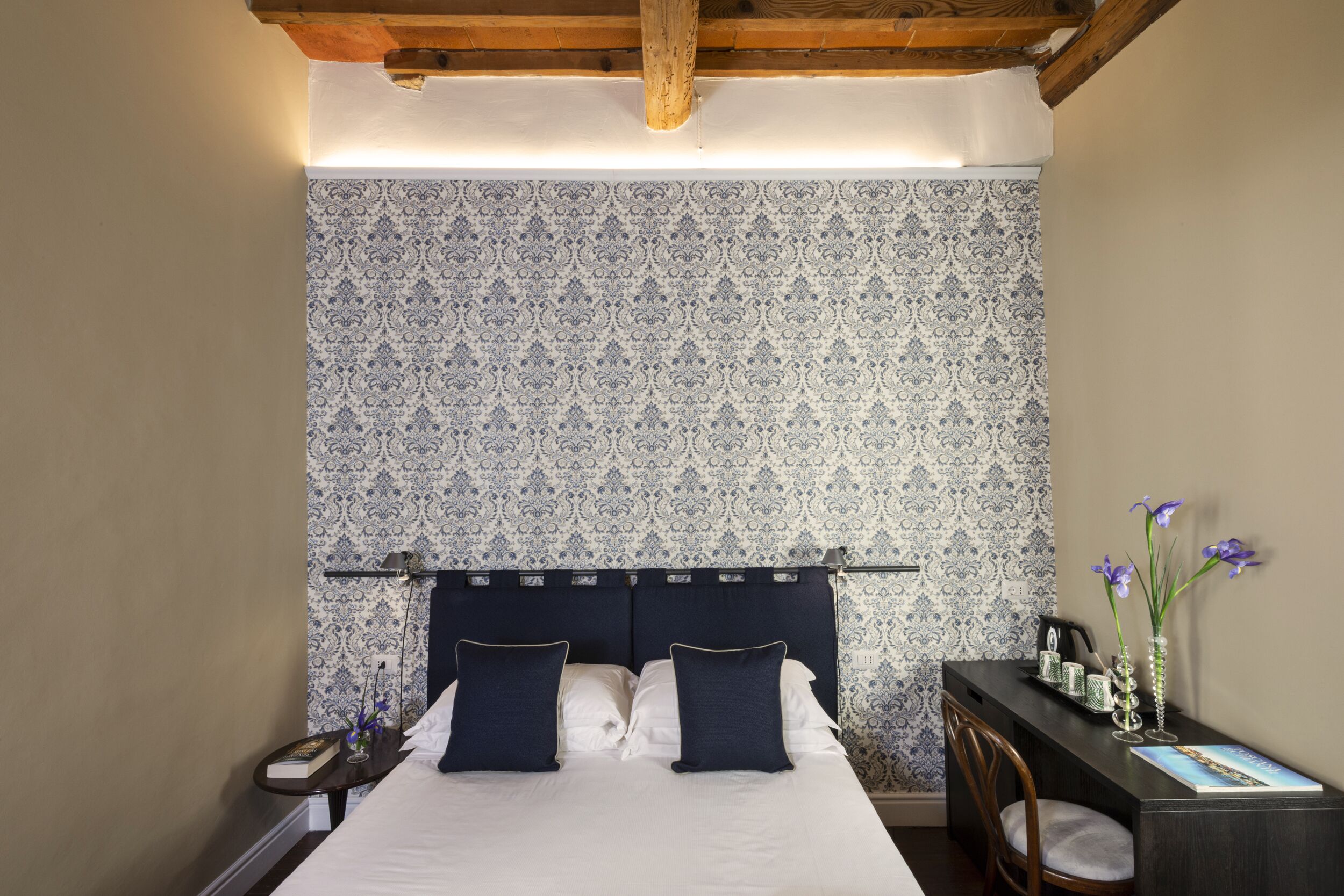 A bedroom with a patterned wallpaper, a bed with navy pillows, a bedside table with a book and a vase, and a desk with a chair and a vase.